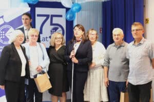 Osiem osób stoi razem na tle baneru jubileuszowego „75 lat” Polskiego Związku Niewidomych, ozdobionego balonami.