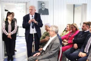 W jasnym pomieszczeniu stoi mężczyzna w granatowym garniturze z białą laską. Po jego lewej stronie stoi kobieta w okularach, a po prawej siedzą inne osoby, w tym Justyna Spryszak. Na ścianie wisi czarno-biały portret mężczyzny.