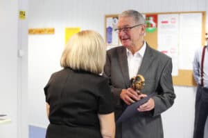 Kobieta w czarnej sukience wręcza dyplom i statuetkę w kształcie uścisku dłoni uśmiechniętemu mężczyźnie w okularach i szarym garniturze. W tle wisi tablica z dokumentami.