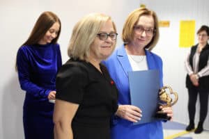 Uśmiechnięta Małgorzata Romanko trzyma niebieską teczkę oraz mosiężną statuetkę w kształcie uścisku dłoni. Obok stoi blondynka w czarnej sukience, a w tle kobieta w chabrowej sukience.