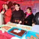 Pięć kobiet prezentuje stoisko Koła Gospodyń Wiejskich pod czerwonym namiotem. Na stole przed nimi stoją tace z domowymi wypiekami, a obok duży metalowy termos. Za kobietami wisi baner KGW Okopy Kolonia.