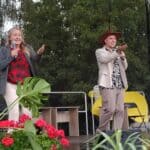 Adam Richaud i jego mama Monika Oleniak-Richaud występują na scenie. Oboje trzymają mikrofony i gestykulują. Scenę zdobią kwiaty i liście.