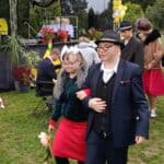 Dwoje uczestników idzie razem po trawniku przed sceną. Mężczyzna ma garnitur i kapelusz, a kobieta zieloną bluzę, czerwoną spódnicę i dużą białą kokardę we włosach. W tle są inni uczestnicy, scena, kwiaty i żółto-białe balony.