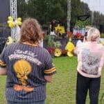 Dwie osoby z publiczności z oddalenia obserwują występ na scenie, którą zdobią kwiaty, rośliny i żółto-białe balony. Obie osoby mają koszulki z nadrukami.