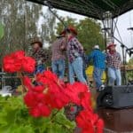Grupa osób w kowbojskich kapeluszach i strojach w stylu western wykonuje układ taneczny na plenerowej scenie. Przed sceną dekoracja z czerwonych pelargonii.