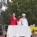 Dwie kobiety odgrywają na scenie scenkę z dużym garnkiem. Jedna ma czerwony żakiet, druga strój kucharki z białym fartuchem i czepkiem. W tle są drzewa i żółte balony.