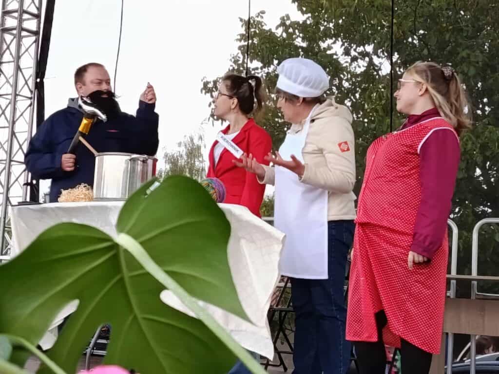 Na scenie, przy stole z dużym garnkiem cztery osoby odgrywają scenkę rodzajową. Mężczyzna w czarnym stroju trzyma duży nóż. Obok niego stoją trzy kobiety: jedna w białym fartuchu i czepku, druga w czerwonym żakiecie, a trzecia w czerwonym fartuchu w białe kropki.
