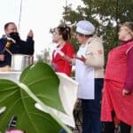 Na scenie, przy stole z dużym garnkiem cztery osoby odgrywają scenkę rodzajową. Mężczyzna w czarnym stroju trzyma duży nóż. Obok niego stoją trzy kobiety: jedna w białym fartuchu i czepku, druga w czerwonym żakiecie, a trzecia w czerwonym fartuchu w białe kropki.