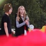 Dwoje młodych uczestników występuje na scenie. Dziewczyna w czarnej kamizelce i biało-czarnej bluzce trzyma mikrofon. Obok niej stoi osoba w czarnym stroju i czapce w kształcie głowy dinozaura.