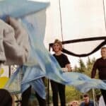 Uczestnicy przedstawienia poruszają dużą błękitną tkaniną, która faluje na scenie. W tle stoją osoby w czarnych strojach, w tym osoba w czapce w kształcie głowy dinozaura.