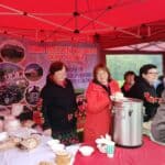 Kilka kobiet prezentuje stoisko Koła Gospodyń Wiejskich Okopy Kolonia pod czerwonym namiotem. Na stole stoją produkty spożywcze i naczynia, a pośrodku - duży metalowy termos z napojem. Za kobietami wisi baner promujący działalność koła.