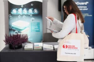 Kobieta przy stoisku konferencyjnym czyta ulotkę. Na ramieniu ma materiałową torbę z logo Hospicjum Dobrego Samarytanina, logo 3. Lubelskiej Konferencji Opieki Paliatywnej i datą 10 października 2026. Na stole leżą broszury i stoi fioletowy wrzos.