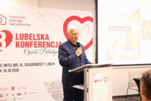 Prelegent w granatowym garniturze przemawia do mikrofonu za mównicą. Po jego lewej stronie stoi ścianka z napisem „3. LUBELSKA KONFERENCJA OPIEKI PALIATYWNEJ” i danymi wydarzenia.