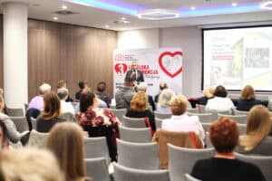 Prelegent w garniturze przemawia na tle ścianki z logo konferencji i dużego ekranu. Przed nim, w rzędach krzeseł, siedzą uczestnicy. Widać ich głowy i plecy.