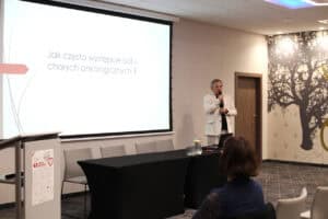 Mężczyzna w jasnej marynarce przemawia do mikrofonu na tle ekranu. Na ekranie widnieje pytanie: „Jak często występuje ból u chorych onkologicznych?”. Przed mężczyzną znajduje się ciemny stół konferencyjny.