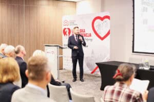 Widok z widowni: mężczyzna w ciemnym garniturze przemawia przy mównicy z logotypami partnerów. Za nim stoi biała ścianka z dużym czerwonym sercem, logo Hospicjum Dobrego Samarytanina i napisem „3. LUBELSKA KONFERENCJA OPIEKI PALIATYWNEJ”.