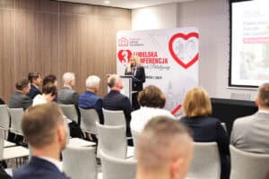 Anna Augustyniak przemawia z mównicy do uczestników na widowni. Za nią stoi biała ścianka z czerwonym sercem, logo Hospicjum Dobrego Samarytanina i napisem „3. LUBELSKA KONFERENCJA OPIEKI PALIATYWNEJ”.