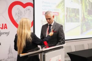 Anna Augustyniak przypina medal na wstążce do klapy marynarki mężczyzny w szarym garniturze. W tle widać białą ściankę z czerwonym sercem i fragment ekranu projekcyjnego.