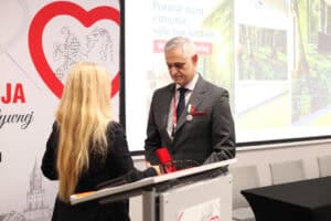 Mężczyzna w ciemnym garniturze patrzy na medal na biało-zielono-czerwonej wstążce, który ma przy klapie marynarki. Przed nim stoi Anna Augustyniak.