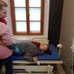 Dwie osoby w Komfortce, czyli pokoju higieniczno-sanitarnym dla dorosłych osób z niepełnosprawnościami i ich opiekunów. Jedna osoba leży na regulowanej leżance, a druga stoi obok podnośnika dla osób o ograniczonej mobilności.