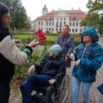Grupa osób stoi wokół krzewu czerwonej róży. W tle widać pałac.
