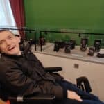 Mężczyzna na wózku inwalidzkim siedzi obok szklanej gabloty z ekspozycją zabytkowych aparatów i akcesoriów fotograficznych.