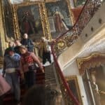 Grupa osób schodzi po schodach. Na ścianach wiszą duże obrazy w złotych ramach.