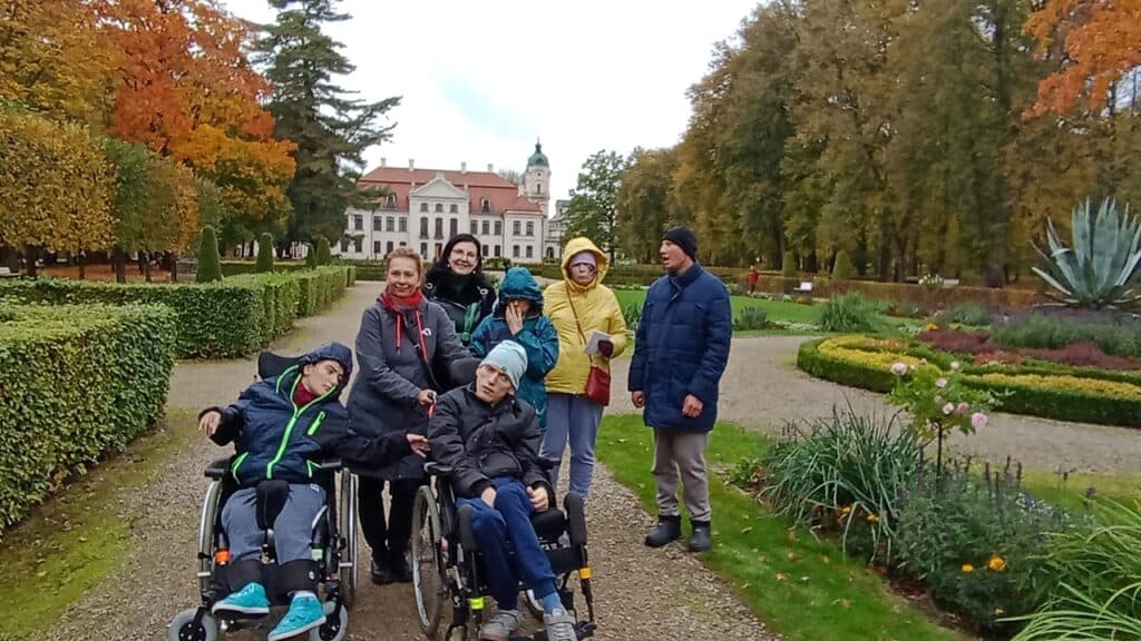 Uczestnicy wycieczki, w tym dwie osoby na wózkach inwalidzkich, pozują w jesiennym ogrodzie pałacowym.