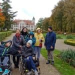 Uczestnicy wycieczki, w tym dwie osoby na wózkach inwalidzkich, pozują w jesiennym ogrodzie pałacowym.