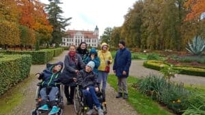 Uczestnicy wycieczki, w tym dwie osoby na wózkach inwalidzkich, pozują w jesiennym ogrodzie pałacowym.