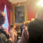 Grupa osób zwiedza komnaty pałacowe. Na ścianie wisi duży obraz w złotej ramie.