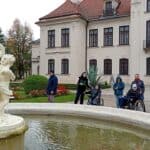 Uczestnicy wycieczki, w tym dwie osoby na wózkach inwalidzkich, stoją między wejściem do Pałacu w Kozłówce a dużą, białą fontanną z rzeźbami postaci.