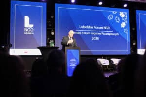 Jarosław Stawiarski przemawia za mównicą z logo Lubelskiego Forum NGO 2026. Za nim znajduje się duży niebieski ekran z grafiką puzzli, a przed sceną siedzi publiczność.