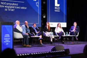 Sześć osób uczestniczy w panelu dyskusyjnym na scenie oświetlonej fioletowym światłem. Za nimi znajduje się ekran z napisami.