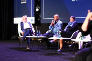Trzech panelistów siedzi na scenie. Mężczyzna w jasnoniebieskiej marynarce przemawia do mikrofonu, a obok niego siedzą kobieta w czarnej sukience i mężczyzna w białej koszuli. Na stolikach stoją butelki z wodą i szklanki.