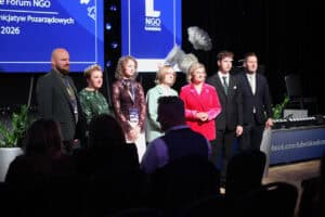 Siedem elegancko ubranych osób stoi obok siebie na scenie i patrzy w stronę publiczności. Za nimi znajduje się niebieski ekran z logo „NGO lubelskie” i napisem „2026”. Scenę oświetlają reflektory.
