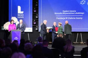 Podczas Gali III Sektora 2026 mężczyzna w garniturze wręcza bukiet kwiatów kobiecie w zielonej sukience. Na ekranie za nimi widnieje napis „Dyplom Uznania Marszałka Województwa Lubelskiego - Fundacja W Zielone Gramy”. Przed sceną siedzi publiczność.