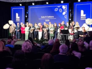 Na scenie stoi grupa osób nagrodzonych podczas Gali III Sektora 2026. Po obu stronach sceny znajdują się duże dekoracyjne kwiaty, a za uczestnikami niebieski ekran multimedialny.