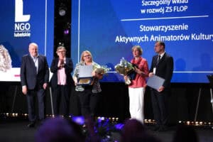 Pięć osób stoi na scenie podczas wręczania wyróżnień. Laureaci trzymają statuetki, pamiątkowe dyplomy w etui i bukiety kwiatów.