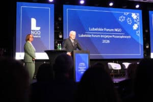 Ryszard Szczygieł przemawia za mównicą podczas Lubelskiego Forum NGO 2026. Obok niego stoi Małgorzata Romanko w zielonym garniturze. Za nimi znajduje się niebieski ekran multimedialny.