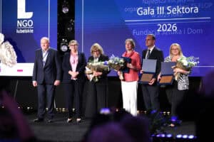 Sześć osób stoi na scenie i prezentuje dyplomy, pamiątkowe tablice z wyróżnieniem oraz bukiety kwiatów. Na ekranie za nimi widnieje napis „Gala III Sektora 2026”.
