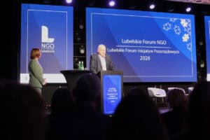 Wojciech Dec przemawia za mównicą podczas Lubelskiego Forum NGO 2026. Obok niego stoi Małgorzata Romanko w zielonym kostiumie. Na ekranie za nimi widnieje napis „Lubelskie Forum NGO”.