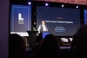 Jacek Gralczyk przemawia za mównicą podczas Lubelskiego Forum NGO. Na ekranie za nim widnieje hasło „NGO to Serce Lokalnej Wspólnoty” oraz dane prelegenta.