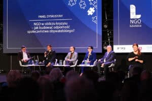 Pięć osób uczestniczy w panelu dyskusyjnym. Nad nimi znajduje się duży niebieski ekran z napisem „NGO w obliczu kryzysu – jak budować odporność społeczeństwa obywatelskiego?”. Po prawej stronie siedzi tłumaczka języka migowego.