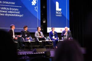 Pięcioro prelegentów siedzi przy stolikach na scenie podczas panelu dyskusyjnego. Za nimi znajduje się niebieski ekran z hasłem przewodnim panelu.