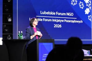 Iwona Kędziera przemawia za mównicą z logo Lubelskiego Forum NGO 2026. Na niebieskim ekranie za nią widnieje napis „Lubelskie Forum NGO Lubelskie Forum Inicjatyw Pozarządowych 2026” oraz grafika puzzli.