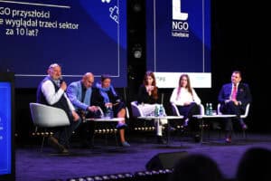 Panel dyskusyjny na scenie z udziałem sześciu osób. Mężczyzna po lewej przemawia do mikrofonu. Na dużym ekranie w tle widnieje napis "NGO przyszłości - jak będzie wyglądał trzeci sektor za 10 lat?".