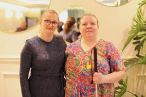 Od lewej: Danuta Bloch w ciemnej, błyszczącej sukience i Ewelina Orzechowska w kolorowej sukience we wzory, z białą laską w dłoni. Obie kobiety się uśmiechają. W tle widać okrągłe lustra oraz roślinę doniczkową.