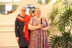 Od lewej: Danuta Bloch w ciemnej sukience i pomarańczowym szalu na ramionach oraz Ewelina Orzechowska w kolorowej sukience we wzory, z białą laską w dłoniach. Obok stoi duża roślina doniczkowa.