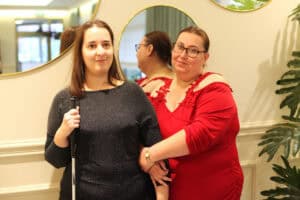 Dwie kobiety stoją obok siebie. Kobieta po lewej ma na sobie ciemną, błyszczącą bluzkę i trzyma białą laskę. Druga kobieta nosi czerwoną sukienkę z odkrytymi ramionami i koronkowym wykończeniem. Kładzie dłoń na przedramieniu towarzyszki.